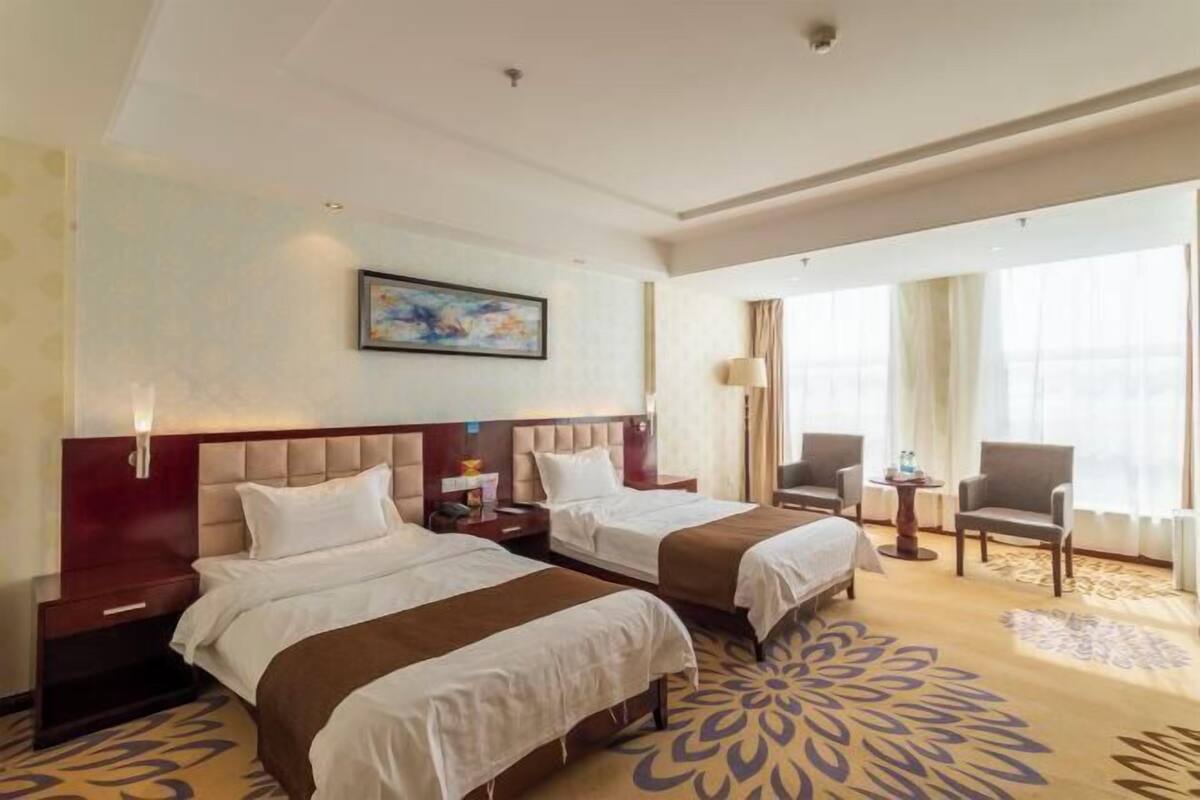 Una habitación de hotel con dos camas, un ventanal con cortinas, una mesita pequeña y una silla.