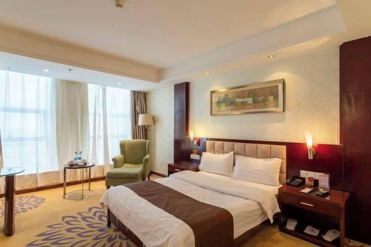 Una habitación de hotel con una cama grande, un sillón verde, una mesita con una lámpara y un cuadro en la pared.