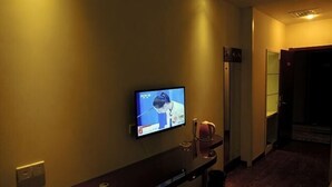 A hotel room with a television mounted on the wall, a wooden bench, a door, and a hallway.