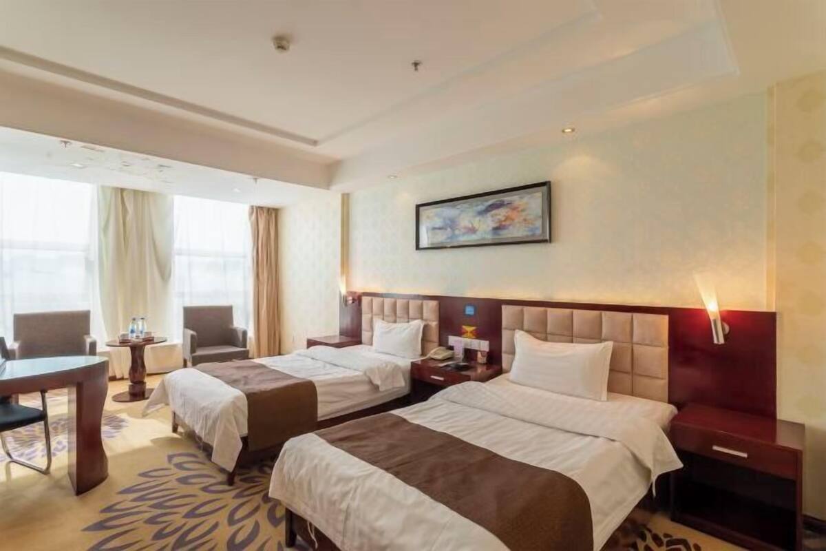 Una habitación de hotel con dos camas, un escritorio, sillas, un cuadro en la pared y una ventana con cortinas.