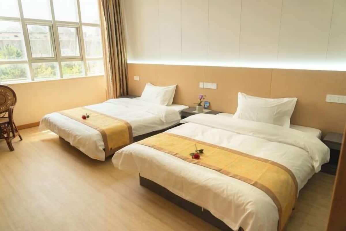 A hotel room with two beds, a wooden floor, a window with curtains, and a nightstand with a lamp.