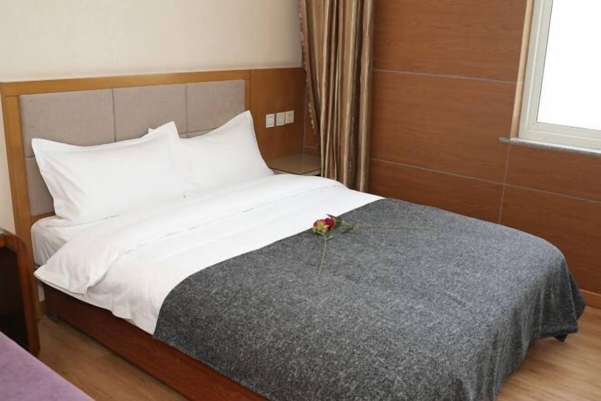 A neatly made bed with white linens and a grey blanket, a single rose on the bed, and a wooden headboard.