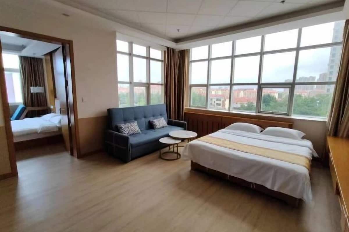 A hotel room with a bed, a sofa, a small table, and large windows.