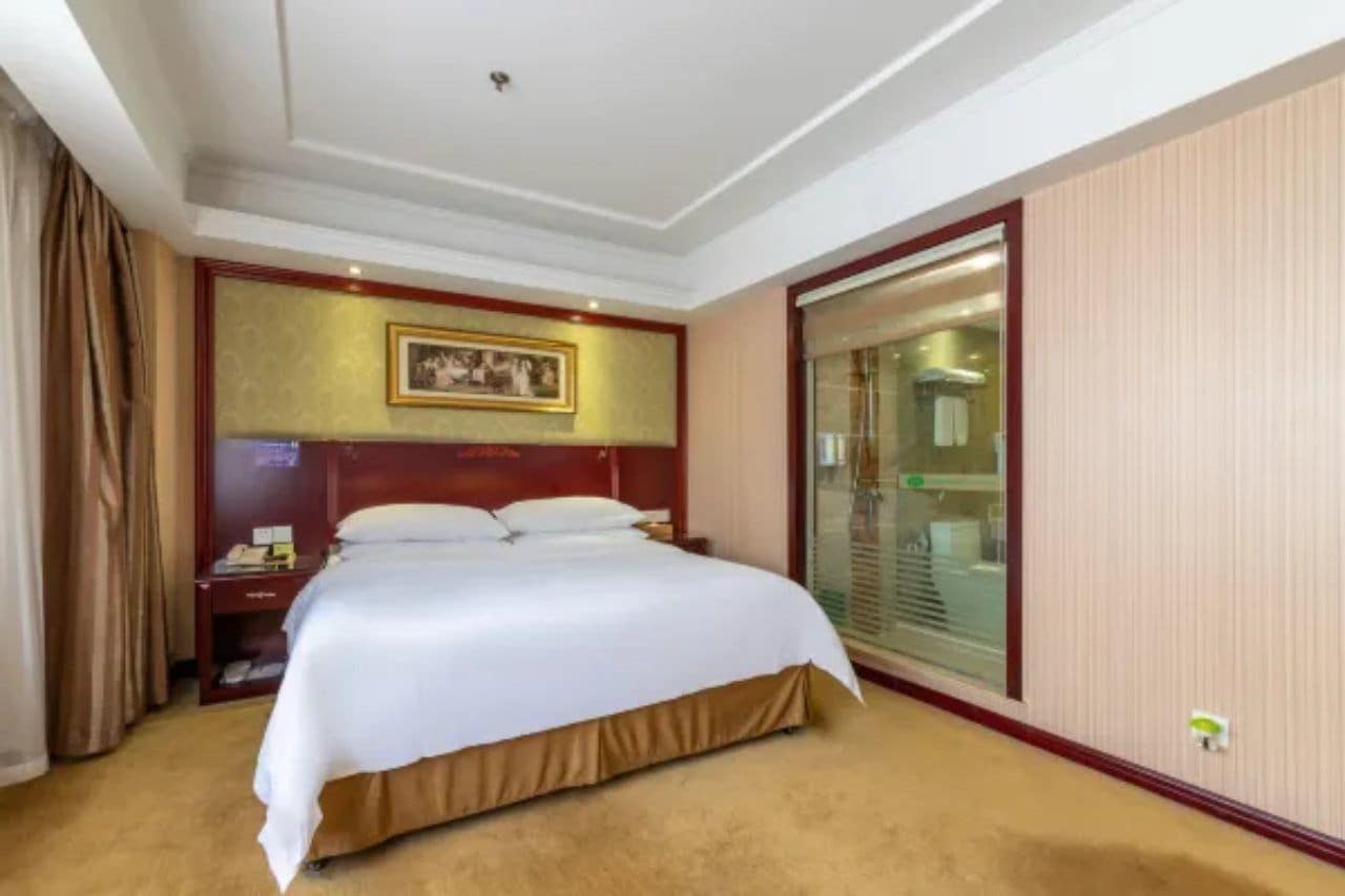 A hotel room with a large bed, a bedside table, a nightstand, a framed picture on the wall, and a sliding glass door leading to a bathroom.