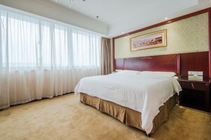 Una habitación de hotel con una cama grande, un cabecero de madera, un cuadro enmarcado en la pared y una ventana con cortinas traslúcidas.