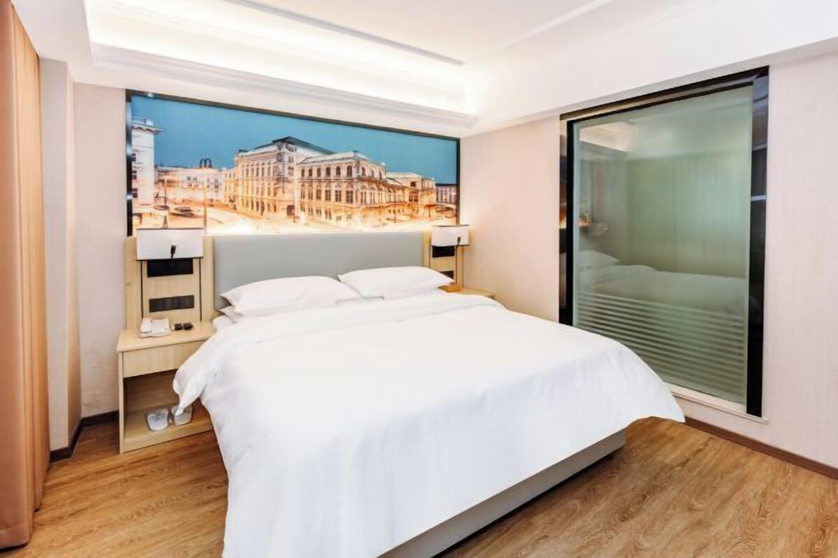 A hotel room with a large bed, a nightstand with a lamp, a wall-mounted TV, and a window with blinds.