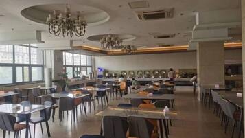 A spacious dining area with multiple tables and chairs, large windows, and a kitchen area in the background.