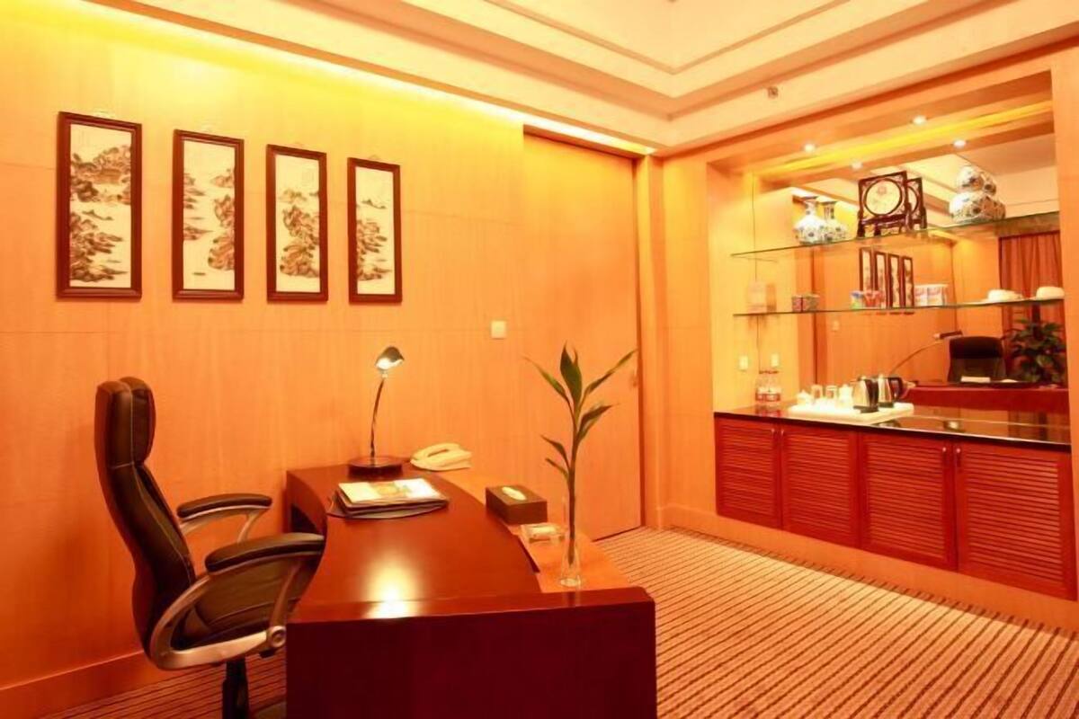 A conference room with a wooden table, a single chair, framed artwork on the wall, and a kitchen area with cabinets and shelves.