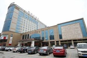 A hotel with a modern facade, a large central entrance, and a parking lot with several cars. - Xin Ci Hotel (Yancheng)