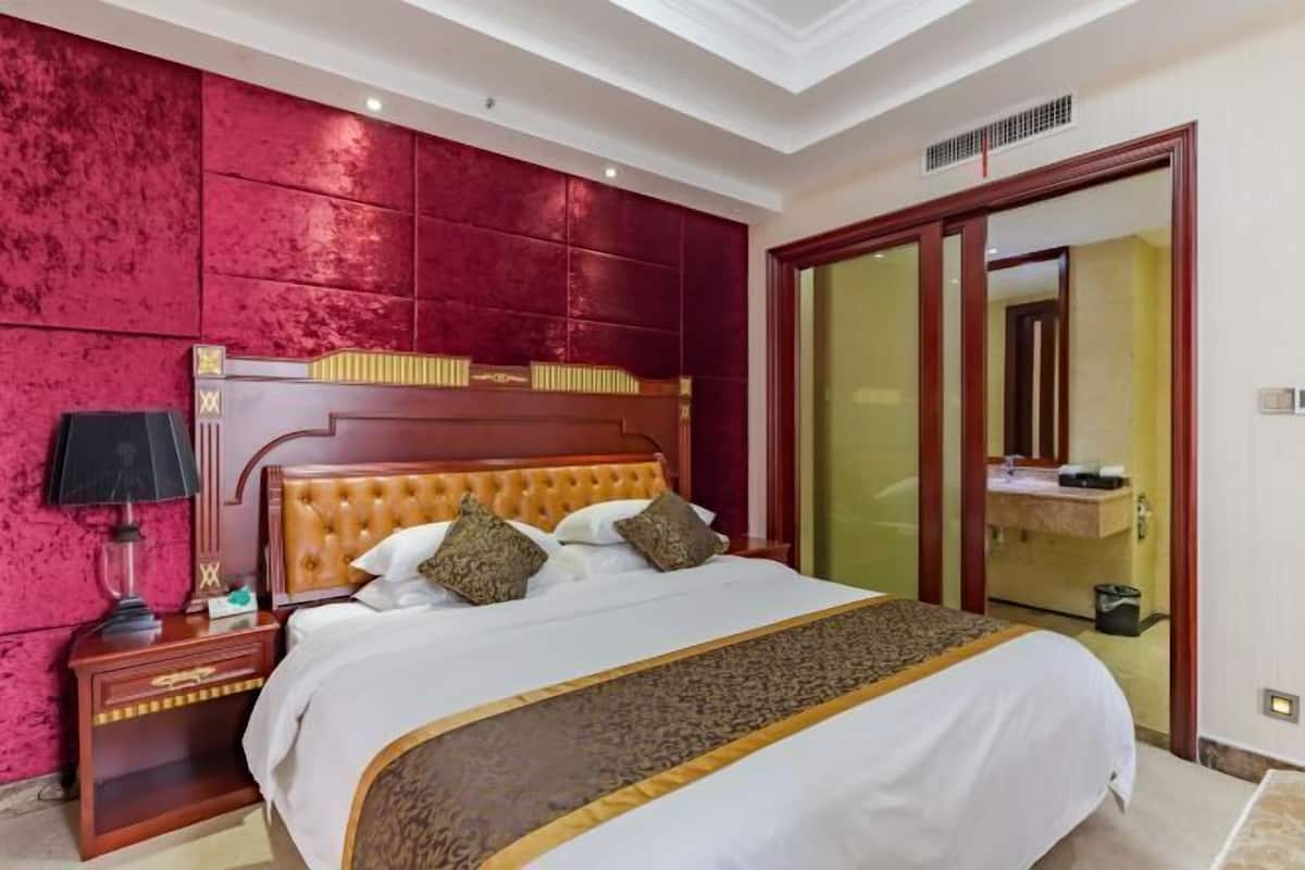 A hotel room with a large bed, a red textured wall, a wooden headboard, a nightstand with a lamp, and a bathroom with a mirror and sink.