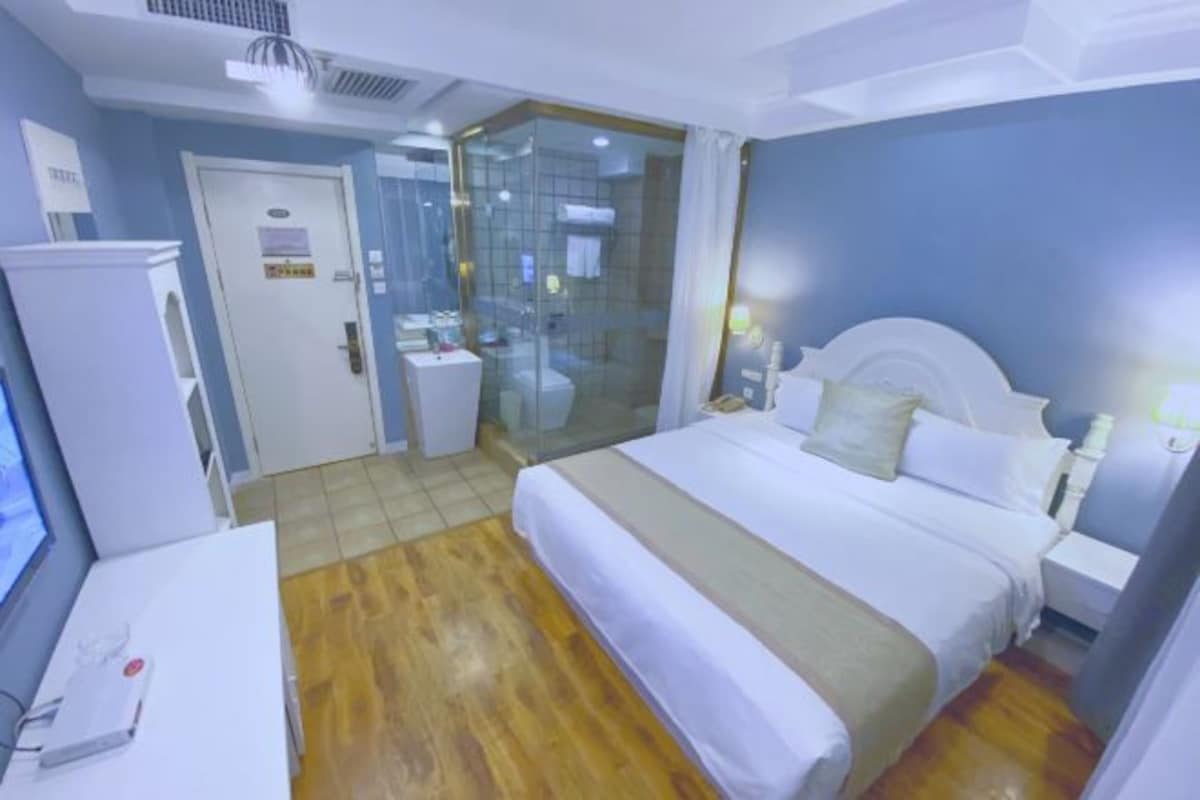 A hotel room with a bed, bedside tables, a desk, a wardrobe, and a glass-enclosed shower area.