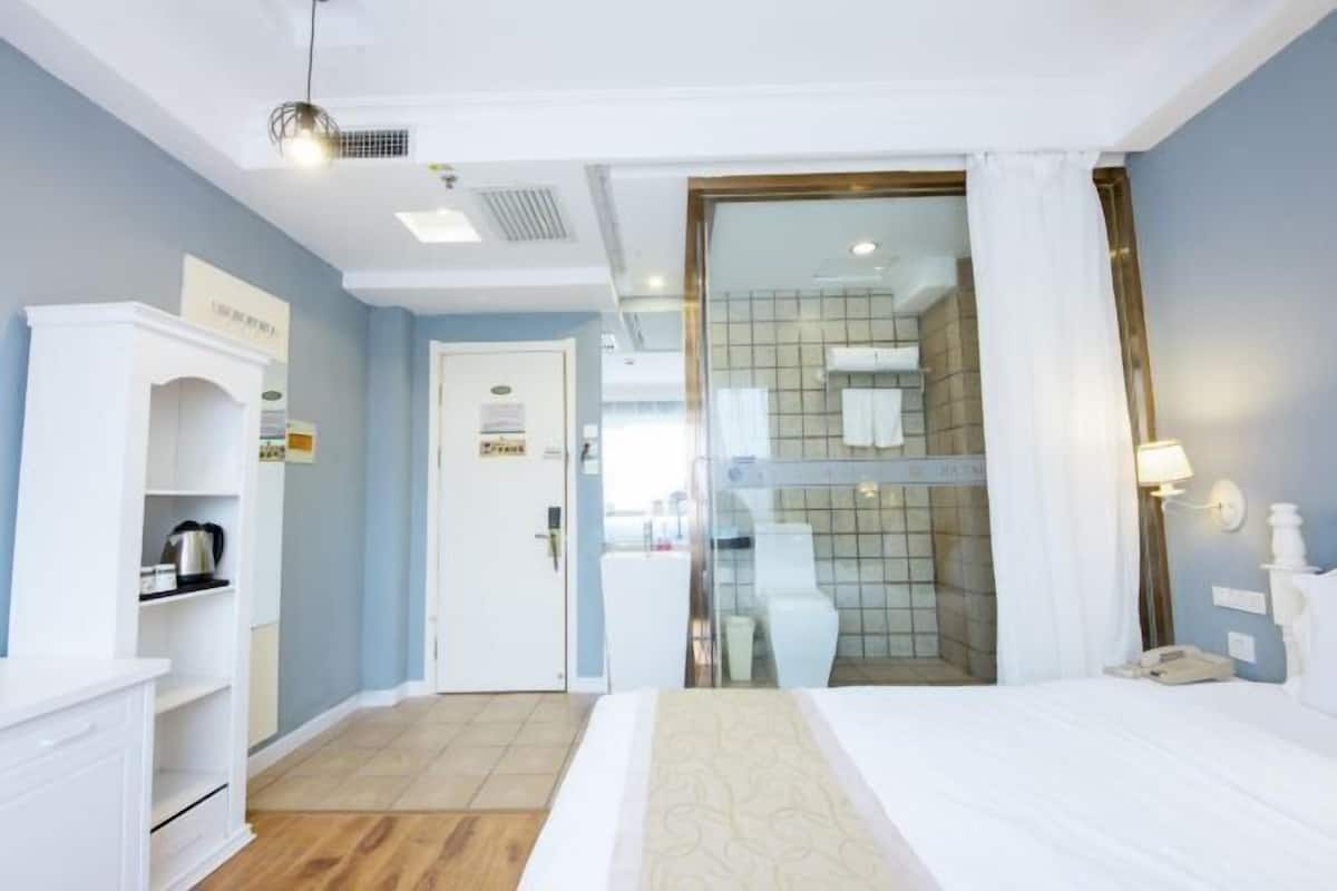 A hotel room with a bed, a wardrobe, a small kitchenette, and a bathroom visible through a glass door.