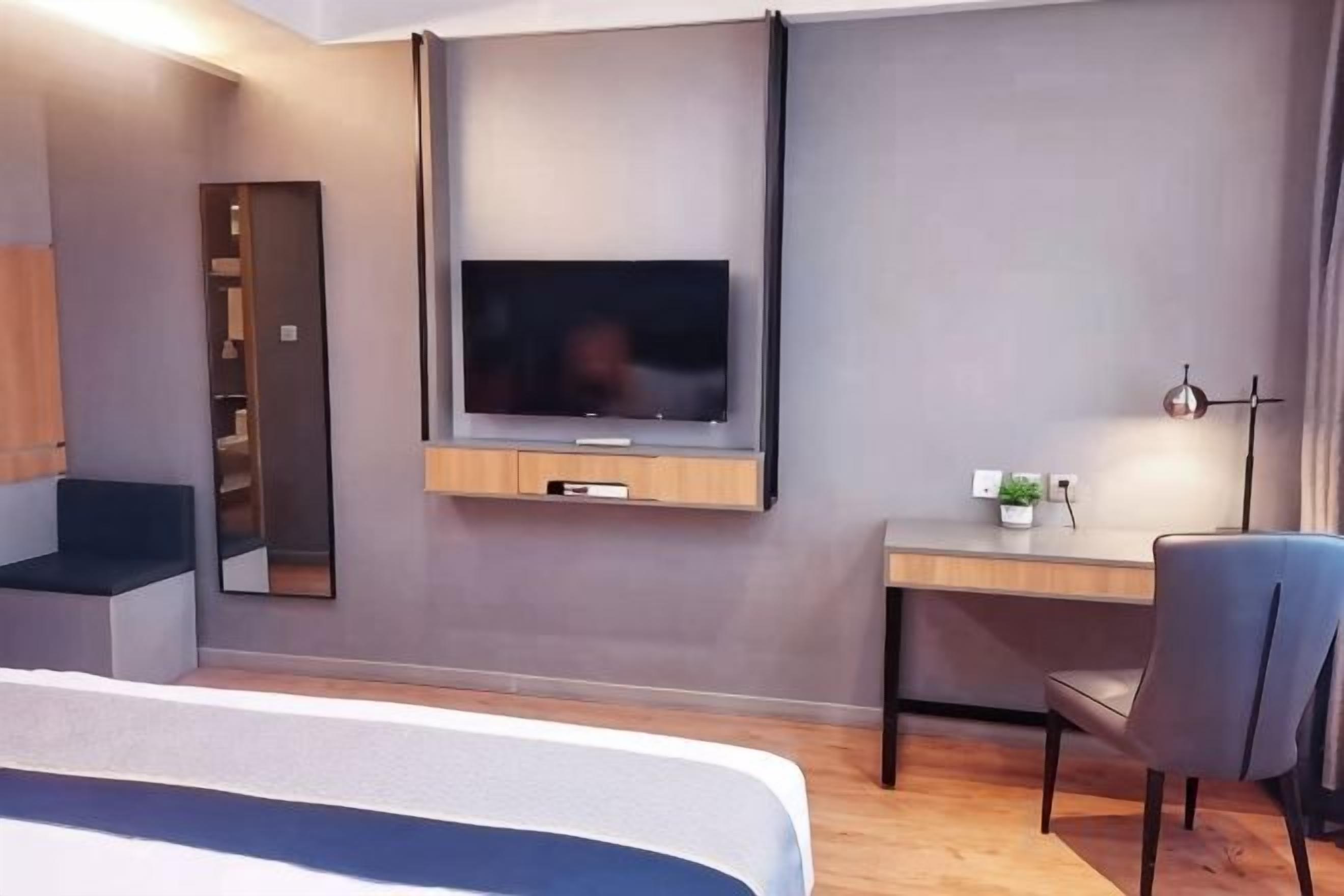 Una habitación de hotel moderna con una cama, un televisor montado en la pared, un escritorio con una silla y una pequeña planta en el escritorio.
