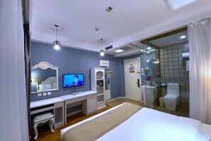 A bedroom with a bed, a desk, a television, a mirror, and a bathroom with a glass shower enclosure. - Jiatai Chain Business Hotel (Zhuanghe High-speed Railway Station) (Zhuanghe)
