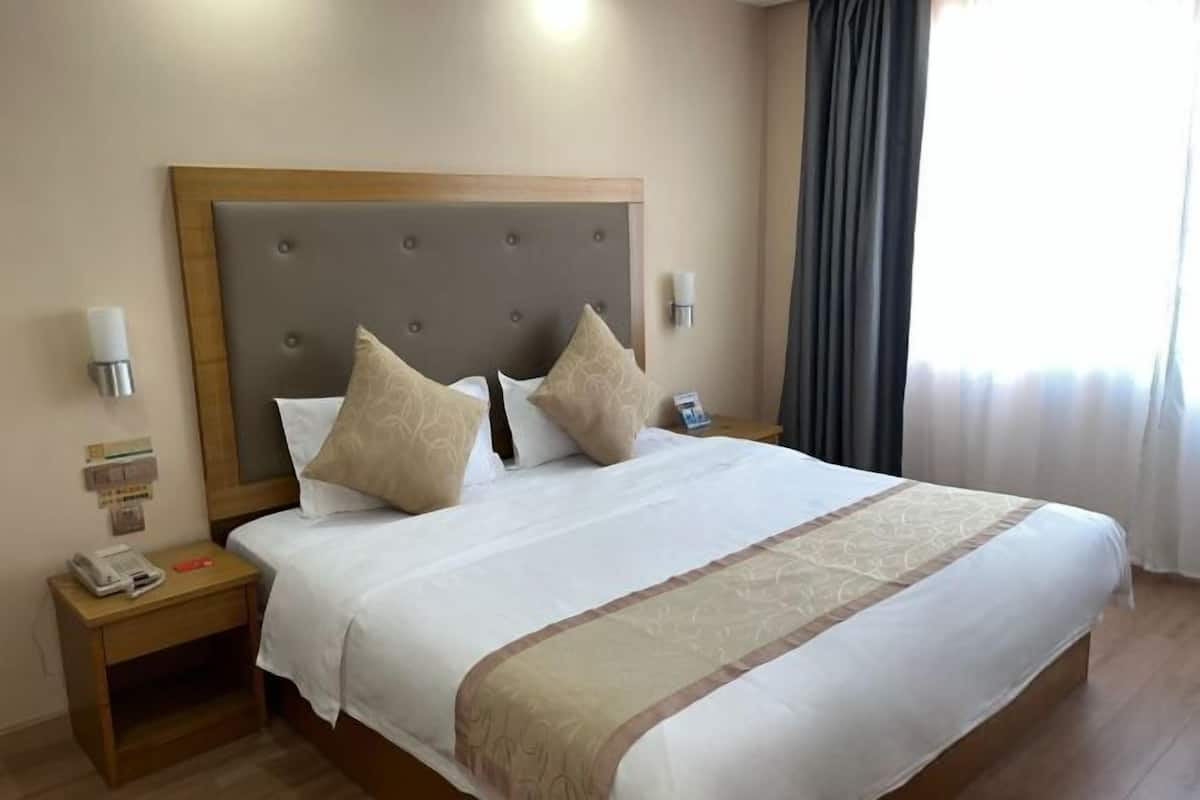 A hotel room with a large bed, a wooden headboard, two bedside lamps, a nightstand with a phone and a red object, and a window with curtains.