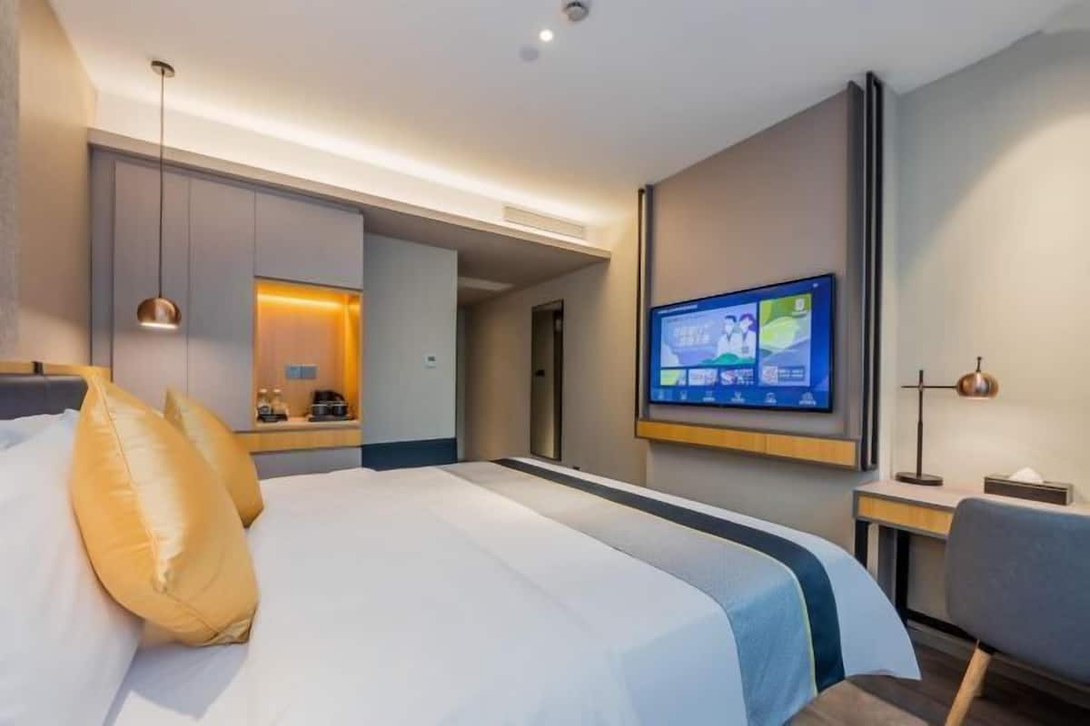 A modern hotel room with a large bed, a flat-screen TV mounted on the wall, and a desk with a lamp.