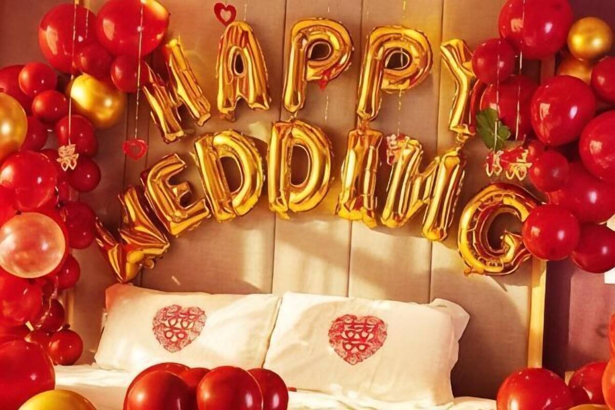 A bed with pillows, a 'HAPPY WEDDING' balloon arch, and red and gold balloons.