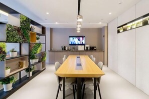 Ein Konferenzraum mit einem langen Tisch, Stühlen, einem Fernseher und einer Küchenzeile inklusive Schränken und Geräten.
