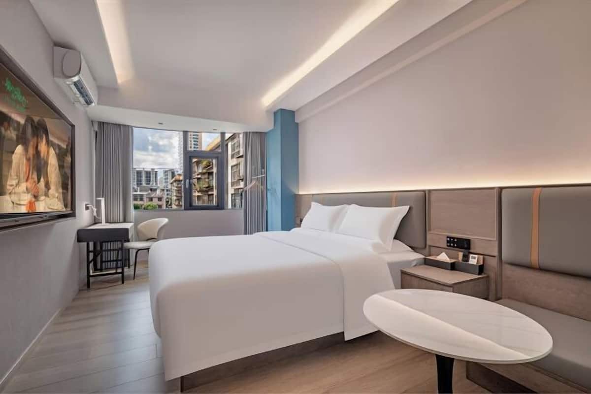 A modern hotel room with a large bed, a desk, a chair, a bedside table, and a view of the cityscape.