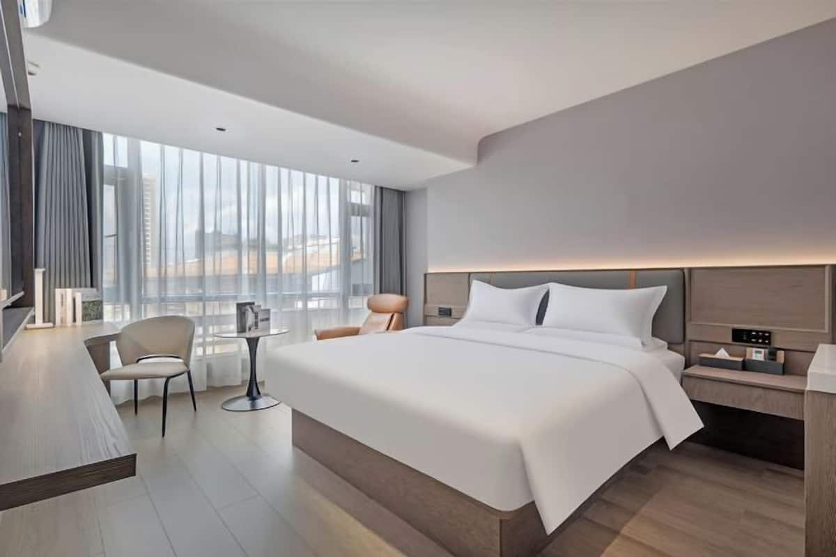 A modern hotel room with a large bed, a desk, a chair, and a view of the city.