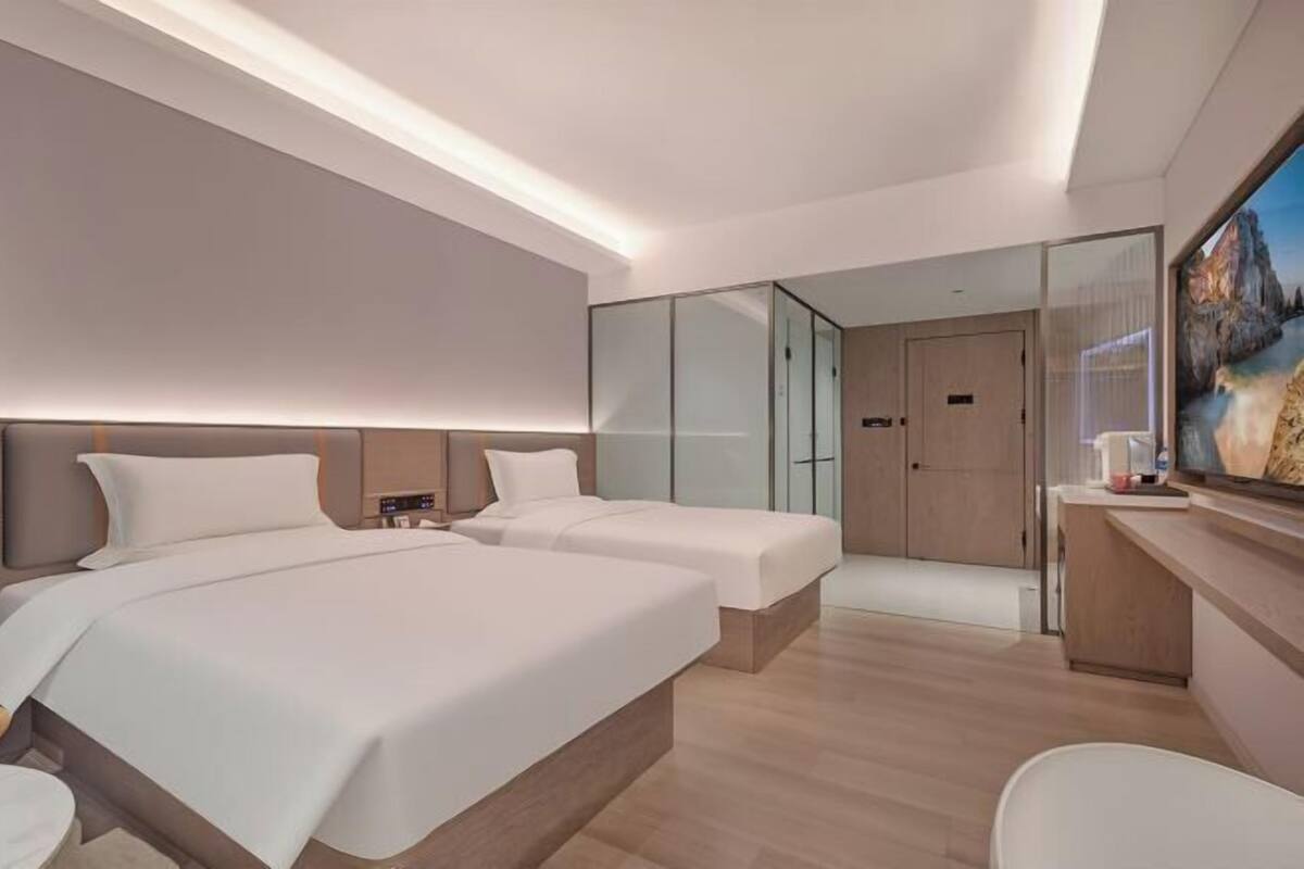 A modern hotel room with two beds, a glass partition, a desk, and a large window.