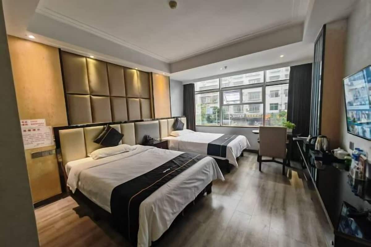 A hotel room with two beds, a desk, a chair, and a large window offering a city view.