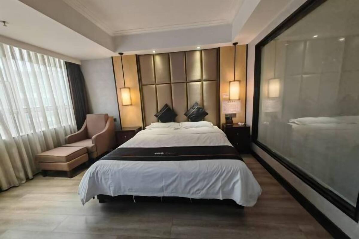 A modern hotel room with a large bed, a chaise lounge, and a large window with curtains.