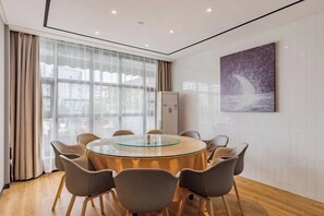 Un comedor moderno con una mesa redonda de vidrio rodeada de sillas, ventanas amplias con cortinas y una obra de arte colgada en la pared.