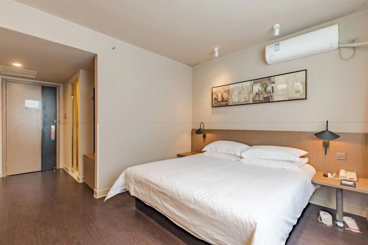 A hotel room with a large bed, a bedside table with a lamp, an air conditioning unit, and a framed picture on the wall.