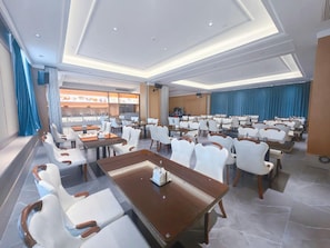 A hotel conference room with multiple tables and chairs, a kitchen area in the background, and a view of the cityscape through large windows. - Vienna Hotels Group Zhuhai (Zhuhai)