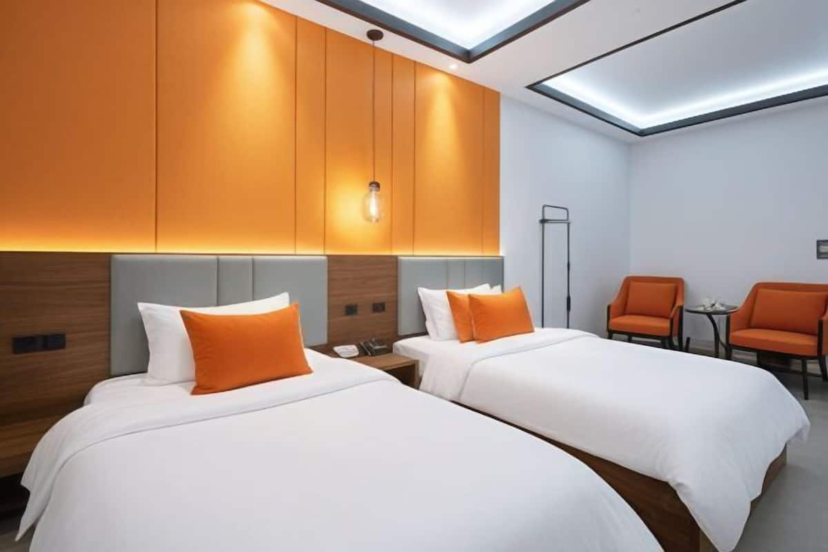 A hotel room with two beds, orange pillows, a small table, and orange chairs.