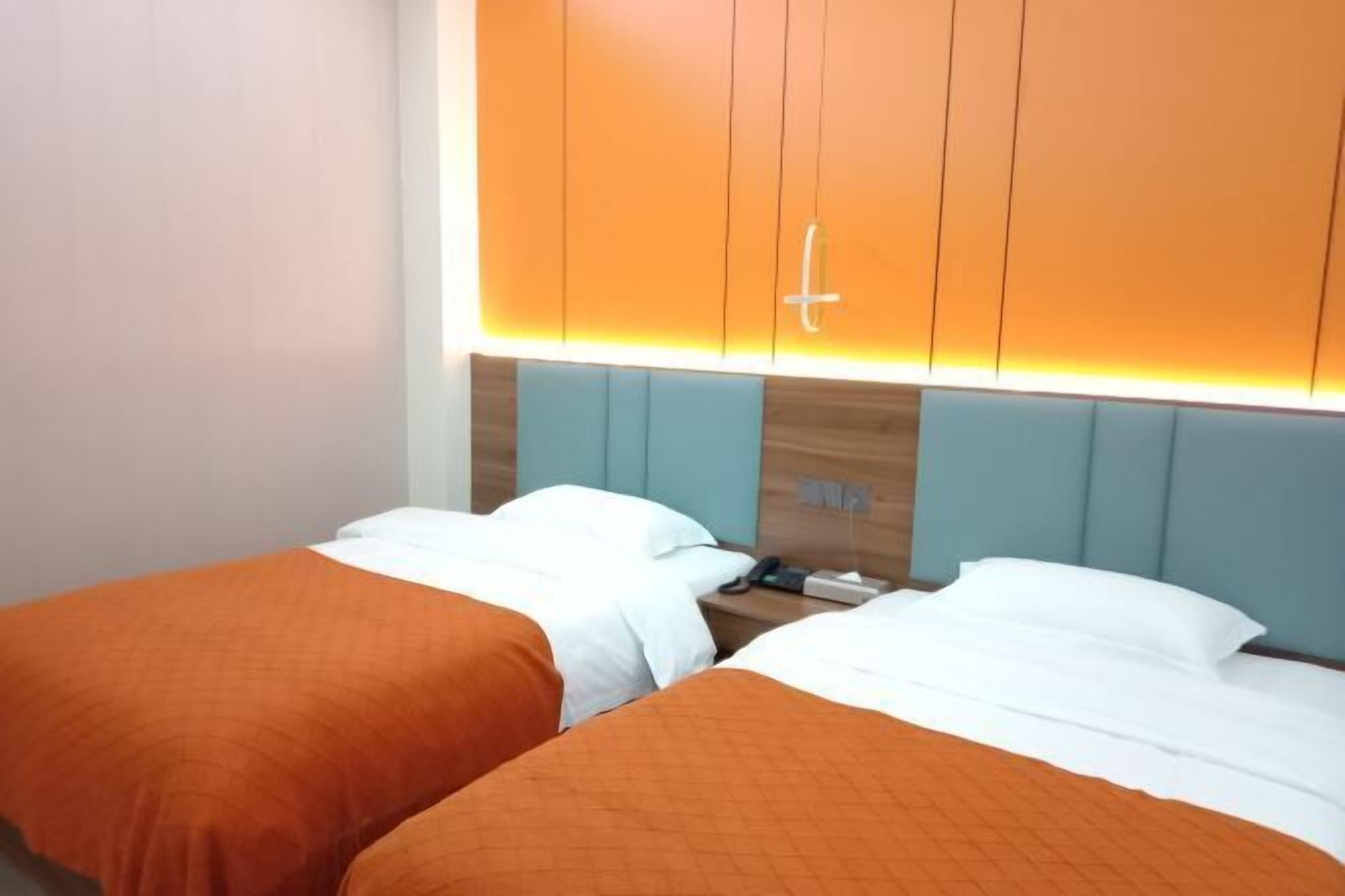 A hotel room with two beds, each with an orange bedspread and white pillows. The headboard is light blue, and there is a wooden panel design on the wall behind the beds.