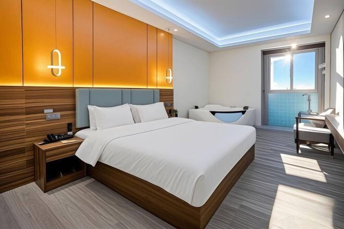 A modern hotel room with a large bed, wooden headboard, built-in wardrobe, bedside table, and a view of the sky through a large window.