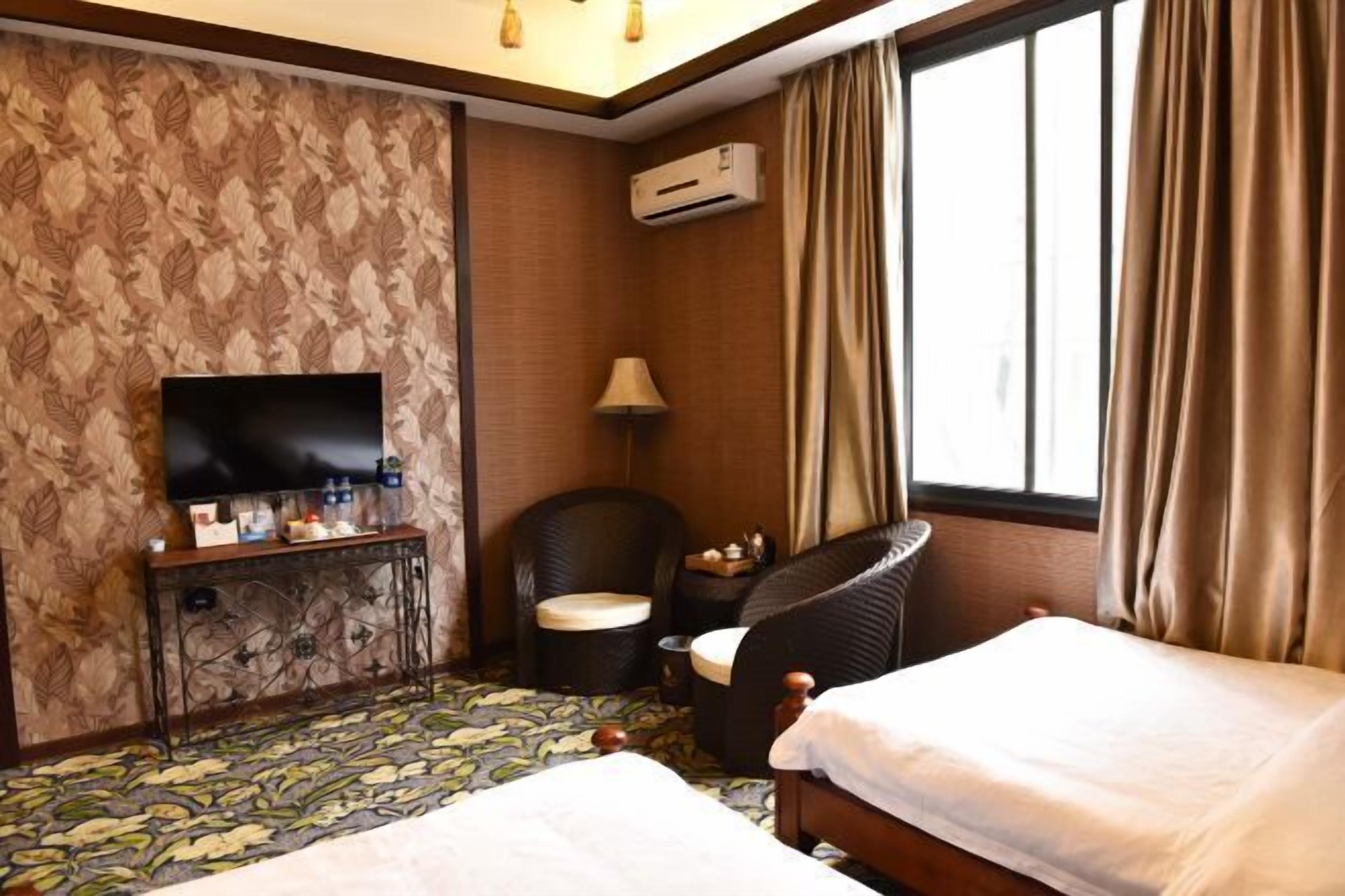 A hotel room with a bed, a TV, a chair, a small table, and an air conditioning unit.