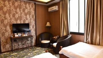 A hotel room with a bed, a TV, a chair, a small table, and an air conditioning unit.