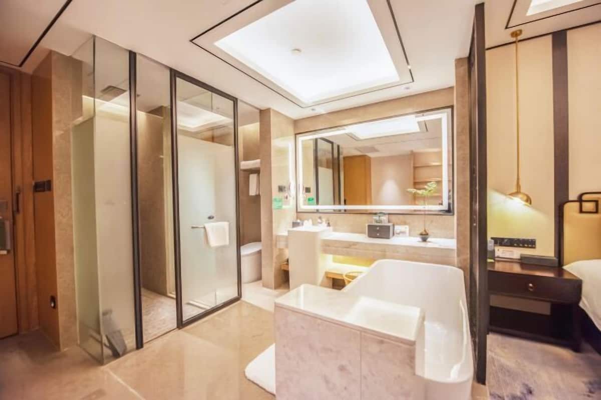 A modern bathroom with a glass shower enclosure, a freestanding bathtub, a vanity with a mirror, and a bedside table with a lamp.