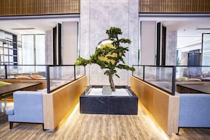 Un espace d’accueil agrémenté d’un arbre bonsai central, d’un comptoir de réception en bois et de rampes en verre.