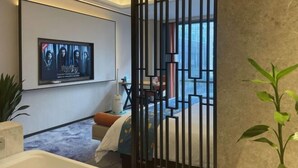 Ein Hotelzimmer mit einem großen an der Wand befestigten Fernseher, einem Bett mit blauen und weißen Kissen und einer blumigen Tagesdecke, einer Topfpflanze und Blick auf die Stadt durch große Fenster.