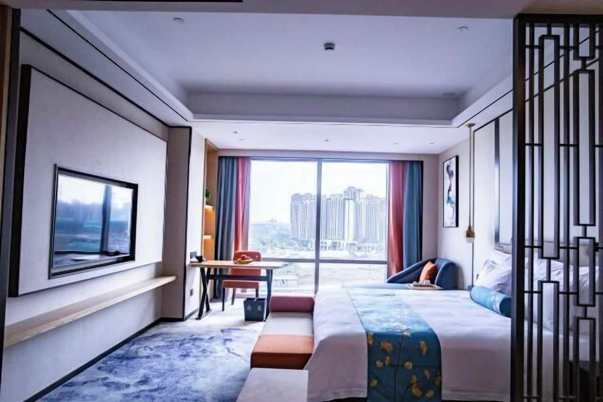 A hotel room with a bed, a desk, a chair, a TV, and a large window with a city view.