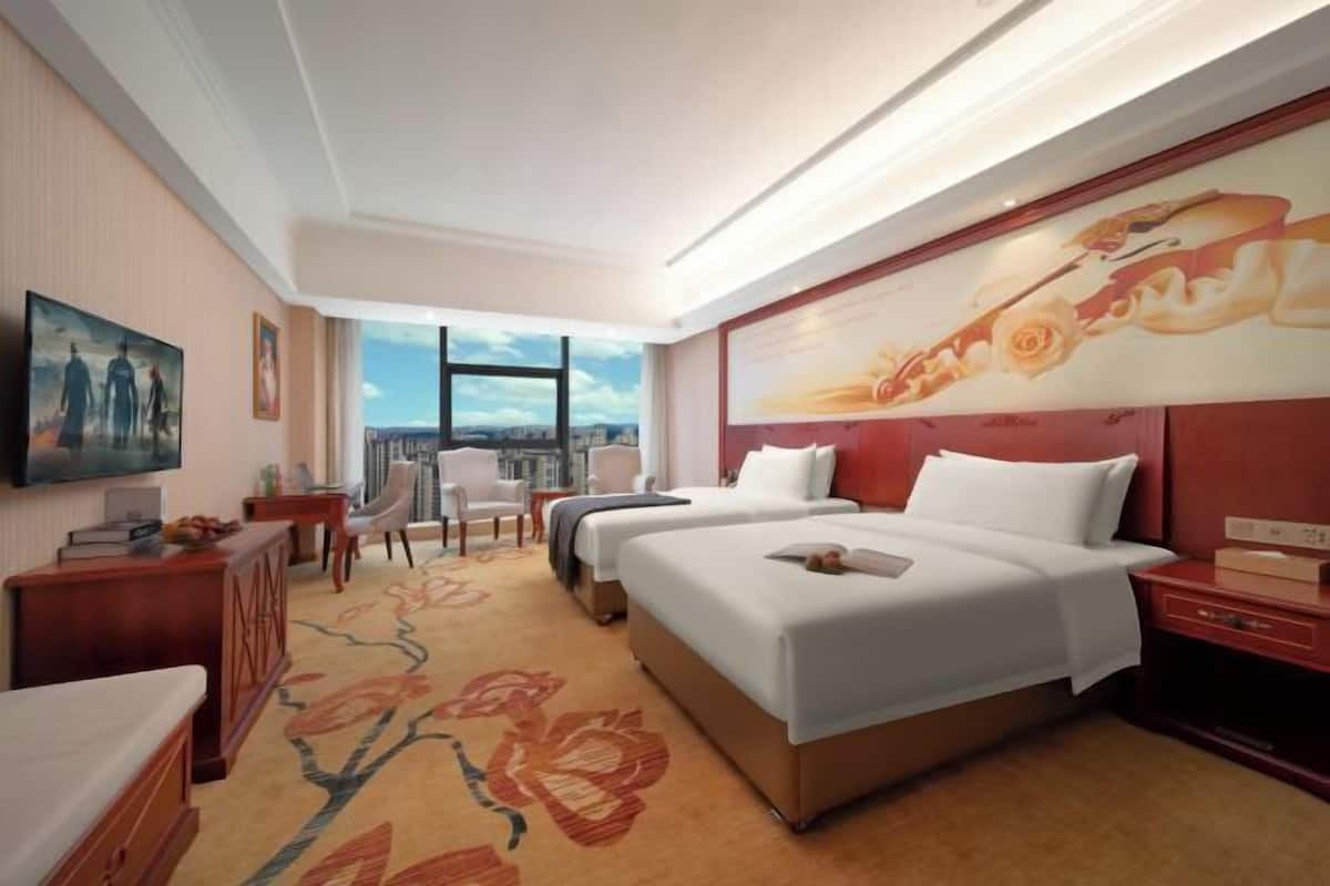 A hotel room with two beds, a large window, a painting on the wall, and a carpet with a floral pattern.