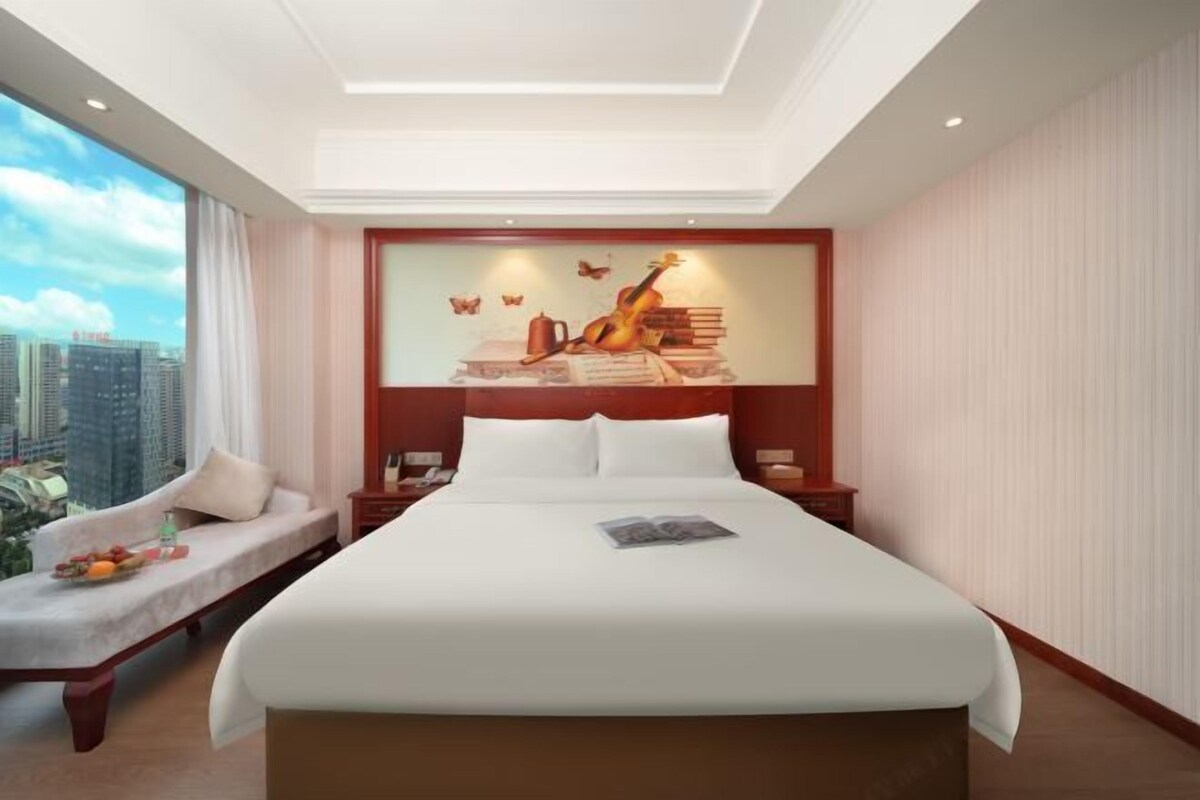 A hotel room with a large bed, a view of the city, a sofa with a tray of fruit, and a mural on the wall.
