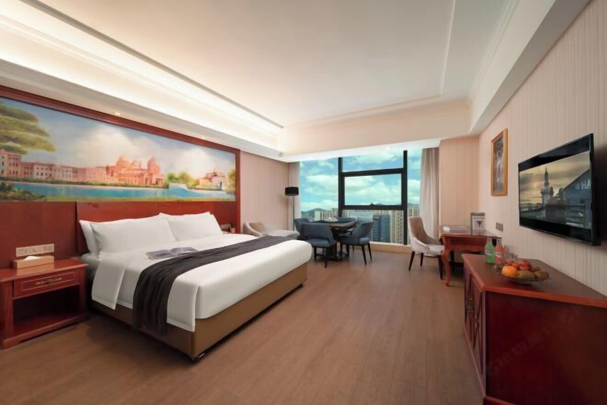 A hotel room with a large bed, a desk, a chair, a TV, and a painting on the wall.