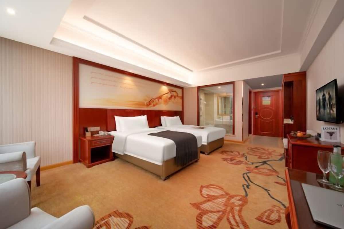 A hotel room with two beds, a desk, a chair, a TV, and a large painting on the wall.