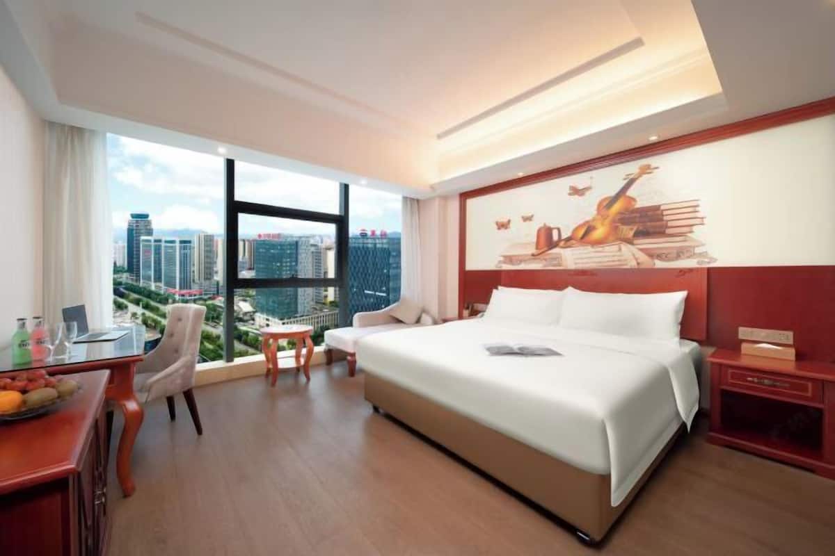 A hotel room with a large bed, a desk with a chair, a small table, and a large window with a city view.