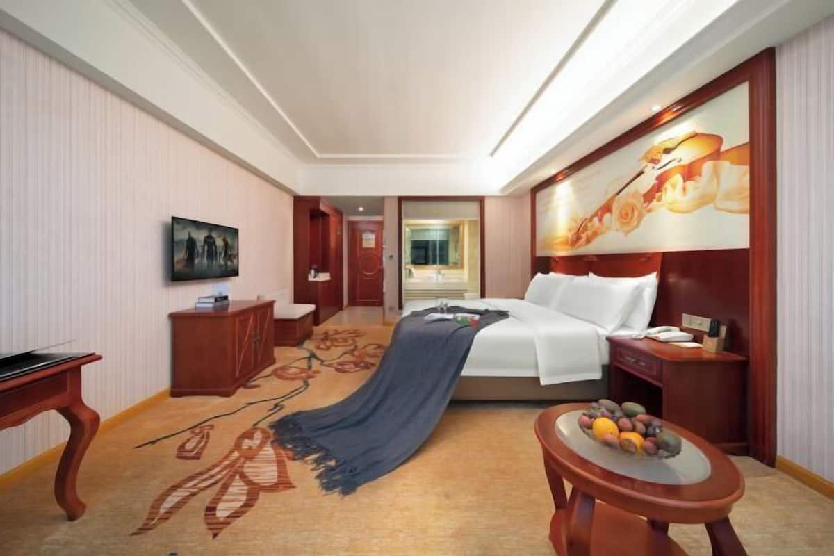 A hotel room with a bed, a desk, a chair, a small table with fruit, and a painting on the wall.