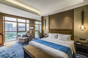 Una habitación de hotel moderna con una cama grande, un escritorio, una silla y vistas de la ciudad a través de amplias ventanas.