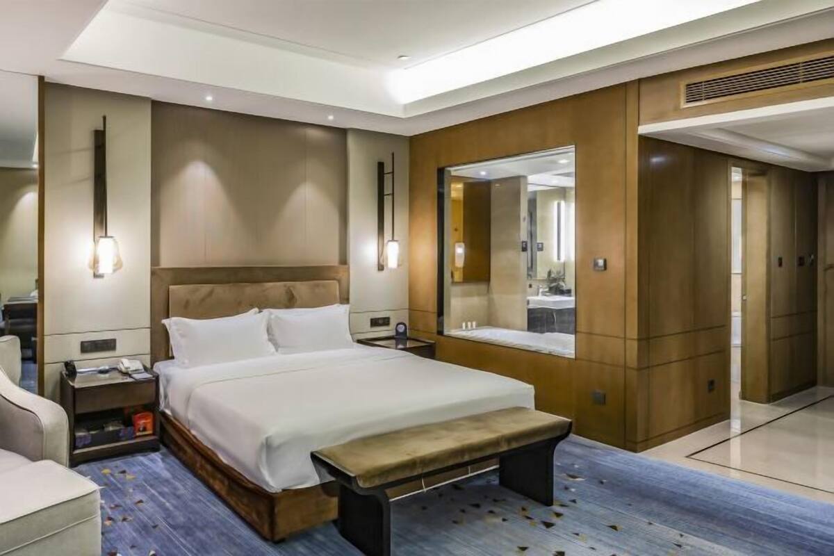 A hotel room with a bed, bedside table, lamp, bench, and a large mirror.