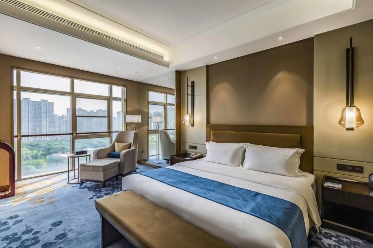 A hotel room with a large bed, a desk, a chair, and a view of the cityscape through large windows.