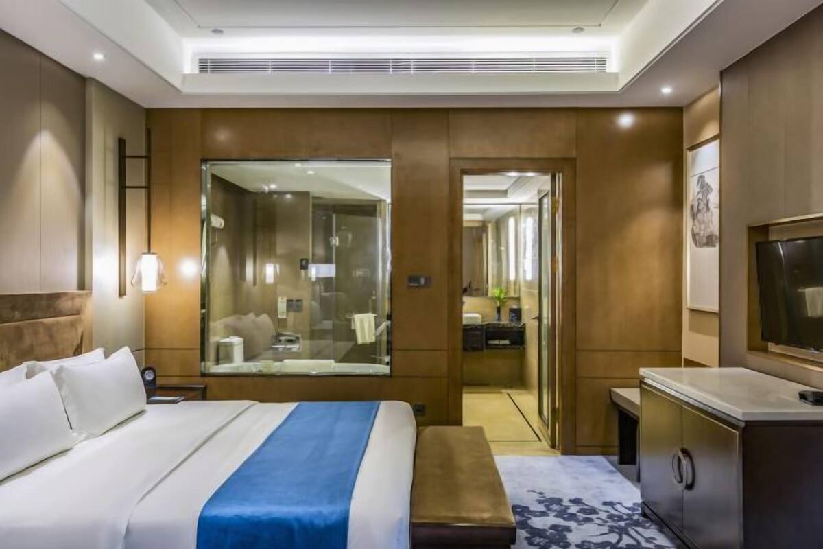 A hotel room with a large bed, a desk, a TV, and a bathroom visible through a glass door.