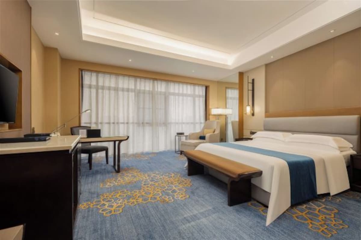 A hotel room with a bed, desk, chair, and a large window.